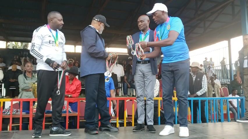 Here is a concise description of the video:

The video shows four men on a stage receiving medals. The man in the blue shirt and white hat is putting medals around the necks of the other men. The man in the black jacket and cap is receiving a medal. The man in the gray suit has a medal around his neck. The man in the white jacket with black stripes also receives a medal. There is a trophy on a stand in the left foreground. Behind the stage, a crowd of people watches.