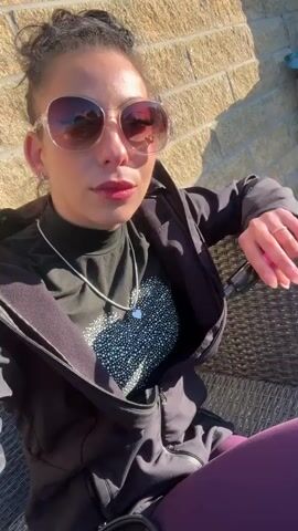 A woman in sunglasses and a black jacket is sitting outdoors in a wicker chair, with a brick wall behind her. She makes a peace sign with her left hand, and is holding a vape pen in her right hand. She sticks her tongue out, then makes a kissing face. She is wearing a black turtleneck shirt with a white heart design, purple leggings, and a silver necklace. Her hair is in a bun. Sunlight is shining on her face and the wall.