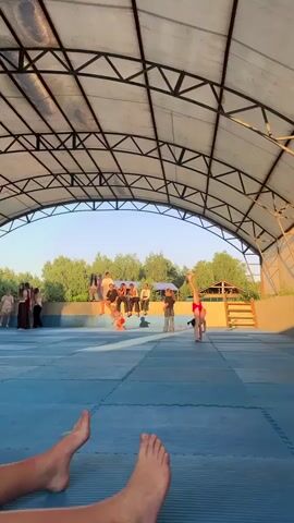 A group of people, predominantly children and young adults, are gathered outdoors under a large, arched, metal-framed canopy. The canopy has a translucent roof, suggesting it provides shade. The ground below is covered with large, blue mats, resembling a gymnastics or martial arts matting.

In the foreground, a pair of bare feet are visible, suggesting the footage is from the perspective of someone lying down or sitting on the ground. In the middle ground, several individuals are engaged in activities. One young person in red shorts and a white top is in the process of performing a cartwheel. Another individual, also in red shorts and a light-colored top, is running across the mats. A group of people are seated on a raised platform behind the matting, observing the activities. They appear to be a mix of ages, with some looking on with interest. Further in the background, trees and vegetation are visible, with a clear blue sky overhead. The lighting suggests it is either early morning or late afternoon, with the sun casting long shadows. The overall atmosphere seems relaxed and recreational, possibly a training session or a casual gathering at an outdoor sports facility. There is no visible text in the media. The location context is Krasnoturansk, Russia.