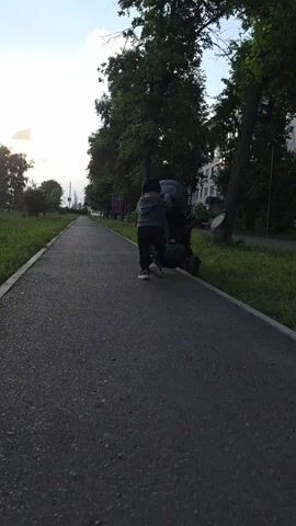 A low-angle shot captures a young child dressed in a green hoodie and dark pants pushing a stroller on a paved walkway. The child is in the process of adjusting something on the stroller, perhaps a bag or a child within, as they are bent over and reaching. The stroller is dark-colored and has a visible green bag attached to it. The walkway appears to be in a park-like setting with lush green grass and mature trees lining the right side. In the background, a white building is partially visible amongst the trees, suggesting a residential area. The sky is light blue with some wispy clouds, indicating it is likely late afternoon or early evening. The overall atmosphere appears calm and serene. No visible text is present in the media file.