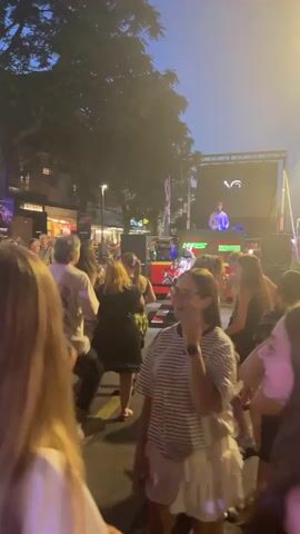 The video shows an outdoor event at dusk or night with a crowd of people dancing. A DJ is visible on a stage with a large screen behind them displaying abstract visuals. The crowd is diverse in age and gender, with individuals dressed casually. Some people are actively dancing, while others are observing or socializing. The environment appears to be a street or public square, with trees and buildings lining the background. The lighting is a mix of stage lights and ambient streetlights, creating a festive atmosphere. The overall scene depicts a lively outdoor party or festival.