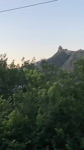 The video shows a view from a moving vehicle, likely a car or train, traveling through a rural landscape in Mtskheta, Georgia. The scene is dominated by lush green trees and vegetation, which blur as the vehicle moves. In the background, a majestic mountain stands tall, crowned with an ancient fortress. The sky is clear with a soft, pale hue, suggesting it is either early morning or late evening. The overall mood is serene and picturesque, capturing the natural beauty of the Georgian countryside. There is no visible text in the video.