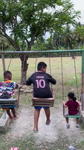 Three children are swinging on a swing set. The child on the left is wearing a multi-colored shirt. The child in the middle is wearing a black shirt with the number 10 and the name MESSI on the back. The child on the right is wearing a maroon shirt. The swing set is green and has three swings. The ground is dirt. There are trees in the background. The sky is cloudy.