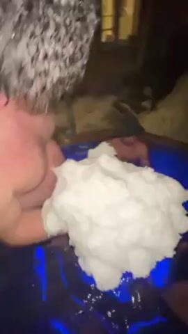 Several men are sitting in what looks like a blue-lit hot tub, and one of the men is holding a large, white fluffy mass of something over the tub. The men appear to be holding the white mass together with their hands. One man is shirtless and is wearing a dark beanie cap. The scene is dimly lit.