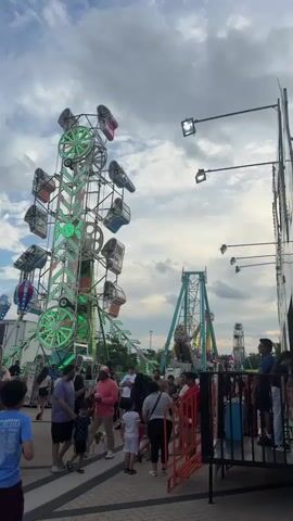 The video captures a lively scene at an amusement park or fair. In the foreground, a tall, green-lit amusement ride with swinging pods is visible. To the right, another amusement ride with a teal A-frame structure and swinging gondolas is in motion. Several people are gathered in the area, with some standing in line and others walking around. The sky above is cloudy, suggesting an overcast day. In the background, other carnival rides, including what appears to be a Ferris wheel, can be seen. The overall atmosphere suggests a busy and enjoyable event.