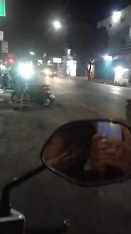 The video shows a street scene at night, viewed from the perspective of someone on a motorbike. In the foreground, the mirror of the motorbike shows a reflection of the rider's face and hands. Further ahead, on the left side of the street, a person is standing next to a parked scooter. Another scooter is also visible. The street is lit by streetlights and the headlights of vehicles. A few other vehicles, including scooters and cars, are present on the road, some moving and some stationary. Buildings are visible on the right side of the street. The overall impression is a busy urban street at night.