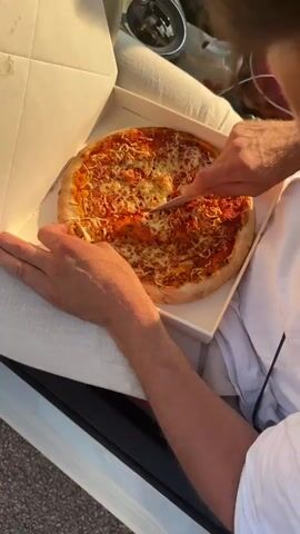 The video begins with a close-up shot of a person's hands cutting a pizza in a white cardboard box. The pizza is topped with tomato sauce and cheese. The camera then pans out to show the pizza box resting inside a car. The interior of the car is visible, including the steering wheel and a part of the dashboard. The scene then shifts to an exterior view of a pizza vending machine labeled "GANG OF PIZZA". The machine is gray and red, with a screen displaying an advertisement for pizza. Text on the machine indicates that it offers gourmet pizza 24/7. The video concludes by showing the pizza box again, with the pizza still being cut.