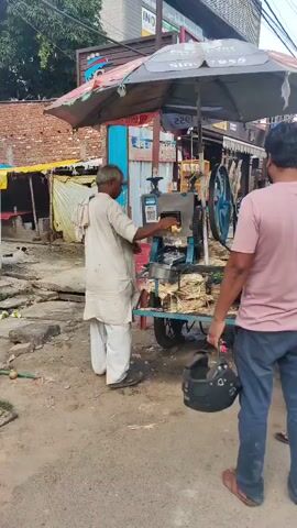 A man in a light-colored kurta and pants stands next to a sugarcane juice machine mounted on a cart. He is operating the machine, which is extracting juice from sugarcane stalks. Several processed sugarcane stalks are visible on the cart. To the right of the machine, another person, partially visible from behind, holds a black helmet. The scene appears to be on a street, with buildings and a canopy visible in the background. The ground is paved and appears somewhat wet.