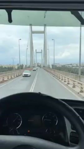 The video shows a driver's perspective from inside the cab of a truck as it crosses a suspension bridge.  The bridge has two parallel lanes of traffic, with a few cars ahead of the truck. The bridge has tall support towers with cables extending down to the road. On either side of the bridge is water, with land and buildings visible in the distance. The day appears overcast. The steering wheel and dashboard of the truck are visible in the foreground.