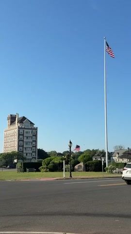 The video shows a tall flagpole with an American flag flying on the right side of the frame. To the left of the flagpole is a multi-story building with a brick exterior and a mansard roof. In front of the building, there are bushes and a lamppost. A second, smaller American flag is visible on the left side of the frame, to the left of the lamppost and in front of the bushes. A road is in the foreground, and the sky is clear blue. There appear to be benches on the grass area. The video is shot from a fixed position, and there is no movement within the scene other than the flags flapping in the wind and what appears to be a bird flying across the sky.