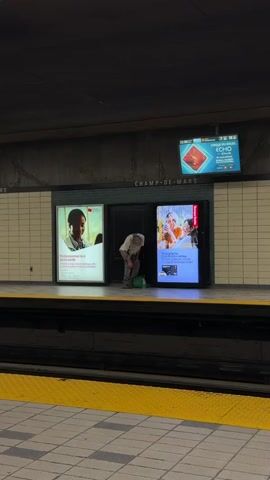 A person with long, light-colored hair, wearing a light-colored top and light-colored pants, is bent over and appears to be looking into a green bucket on the floor. They are standing on a subway platform next to two illuminated advertisements. Above them is a dark sign that says "CHAMP-DE-MARS." In the background, another screen displays a blue and white geometric design. The floor of the platform has light gray and white tiles with a yellow safety strip along the edge. The person remains mostly still throughout the video.