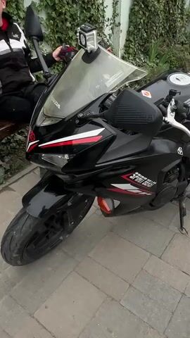 A black Honda CBR motorcycle is parked on a paved surface. The motorcycle has red and white racing stripes. A helmet and gloves are lying on the seat. A person is sitting on a nearby bench. A green vine covers the wall behind the motorcycle.