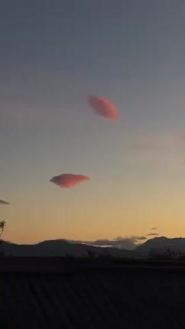 The video captures a picturesque scene at sunset, featuring lenticular clouds in the sky. The clouds are highlighted with a soft pink hue from the setting sun. In the background, a mountain range is visible. The foreground includes a corrugated rooftop. As the video progresses, the camera pans slightly to the right, revealing nearby buildings and a billboard. The sky transitions from a light blue to a warm orange as the sunset intensifies.