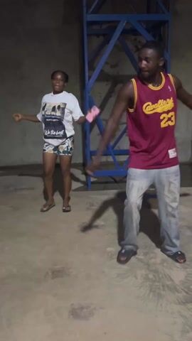 A short video shows a man and a woman dancing in what appears to be a garage or storage area. The woman is wearing a white t-shirt, shorts, and sandals. The man is wearing a basketball jersey with the number 23 on it, jeans, and sandals. Behind them is a blue metal structure, perhaps part of a water tower frame or similar equipment. The floor is concrete and the lighting is dim, giving the impression of an indoor space, likely shot with a handheld mobile phone. The two dancers appear to be enjoying themselves as they move to the music, smiling and interacting with each other.