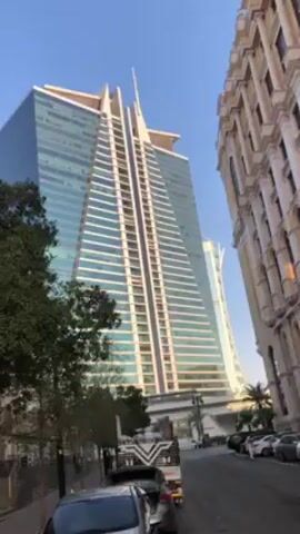 The video presents a view of a street lined with parked cars, flanked by two architecturally distinct buildings. On the left stands a modern glass-clad skyscraper, its shape resembling a triangular prism. To the right, an older, ornate building features light-colored stonework, detailed windows, and classical architectural elements. Small trees line the sidewalk in front of the skyscraper. The sky is clear and blue.