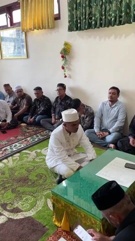 The video shows a group of men sitting on the floor, facing a table covered with a green cloth. Some of the men are wearing white skullcaps, and others are wearing darker headwear. They are all dressed in casual clothing, with some wearing shirts and trousers and others wearing more traditional attire. The room appears to be a meeting room or a hall, with windows and curtains visible in the background. There are framed pictures on the wall. The men are sitting on a carpet, and there are some flowers on a stand in the background. The overall impression is that of a formal gathering or a meeting.