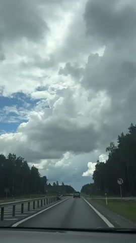 This is a vertical video taken from the perspective of someone in a car driving on a highway. The sky is filled with dramatic, heavy clouds, with patches of blue sky visible. Trees line both sides of the highway. Several cars are driving ahead, with one car in the immediate foreground. There are traffic signs and road barriers visible. The overall mood is overcast and potentially rainy.