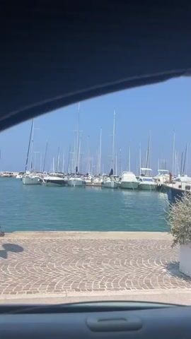 The video shows a view of a marina from inside a vehicle. Several sailboats and motorboats are docked. The water is calm and the sky is clear and blue. There is a paved walkway along the dock with some potted plants and a yellow electrical box. The video pans across the boats, revealing their names and details. One boat is named "Sunshine" and another has the name "Be Quiet III" on its stern. The overall impression is a peaceful and sunny day at a marina.