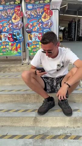 A young man wearing a white t-shirt, black sunglasses, and black sneakers is sitting on concrete steps, looking down at his phone. He is wearing black shorts with a white pattern and a black bracelet on his left wrist. Behind him, a display of ice cream products is visible. Further back, a convenience store with merchandise is open. To the right, another young man, shirtless and wearing patterned shorts, is sitting on the same steps eating from a white plate with a fork. A white plastic bag and a bottle of water are on the steps near him. A woman wearing a black and white patterned bikini top and white shorts is standing and looking at her phone. She is wearing sandals and has her hair pulled up in a bun. She is facing the shirtless man. The steps are made of concrete with yellow and black striped markings. The overall setting appears to be an outdoor area, possibly near a beach or tourist attraction, with a bright, overcast sky.
