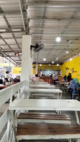 The video shows the interior of a busy restaurant with yellow walls and a corrugated metal ceiling. Rows of white tables and wooden benches are visible in the foreground, while further back, patrons are seated at tables, eating and conversing. A counter area with staff can be seen in the distance. Several fans are mounted on the walls and ceiling to provide ventilation. The overall atmosphere appears lively and casual.