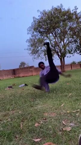 The video shows a person performing a breakdancing move on a grassy lawn. The individual, who appears to be a young man with dark hair, is wearing a light-colored, long-sleeved shirt and dark pants. They start on one knee, then use their arms and legs to spin around on the ground. The setting is an open area with a brick wall in the background. A large tree with green leaves is visible to the right of the person. There are some clothes and shoes lying on the grass nearby. The sky is a pale blue, suggesting it may be either early morning or late afternoon.