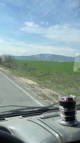 The video shows a driver's perspective while driving on a gray asphalt road. The road is bordered by fields, primarily green with some brown areas. In the distance, there is a mountain range under a bright blue sky with scattered white clouds. Several trees are visible along the roadside, some with white blossoms. The dashboard of the vehicle is gray, and there is a metal and plastic object with coiled wires on the dashboard near the windshield. The vehicle is moving forward, with the road stretching ahead into the distance.
