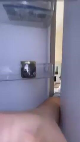 The video opens with a shot inside a refrigerator, focusing on items in the door compartments, including what appears to be yogurt and a beauty face mask. The fridge door is then opened further, revealing a woman in pajamas standing in front of it. She looks directly at the camera, which is apparently mounted inside the refrigerator. The background shows a tiled floor and part of a bed.
