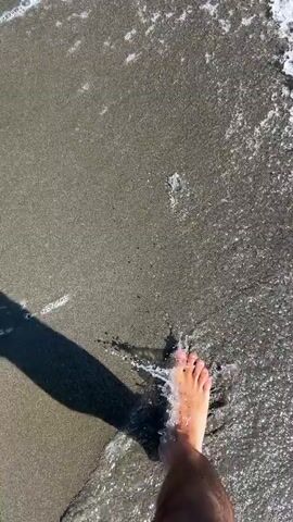 A person's bare foot walks along a sandy beach. The foot is a light skin tone with pink nail polish on the toenails. The foot is stepping in wet sand as a small wave with white foam washes over it. The sun casts a shadow of the person's leg and foot on the sand to the left. The water appears to be dark and the sand is a dark grey color. The video is shot from a first-person perspective looking down at the foot.