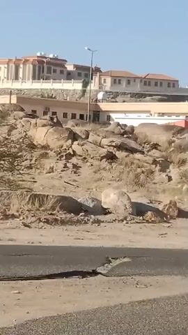 The video shows a street scene with buildings and rocky terrain. In the foreground, a paved road is visible, with some cracks and debris. The middle ground features a rocky, sandy area with sparse vegetation and large boulders. In the background, there are several buildings, including what appears to be a large, light-colored multi-story structure on a hilltop, and other smaller residential or commercial buildings closer to the road. The sky is clear and blue, suggesting a sunny day. The camera appears to be moving, possibly from a vehicle, as indicated by the slight motion blur and the perspective shift. No people or specific actions are clearly discernible.
