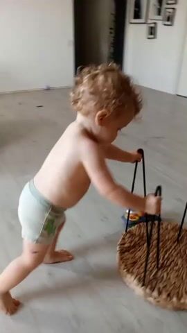 A toddler with curly blonde hair and wearing a diaper is pushing a circular woven basket with black legs across a light-colored floor. The toddler is facing away from the camera and is leaning forward, with their hands on the basket's legs. There are toys on the floor around the basket, including a pink ball and some purple streamers. The background is blurred, but appears to be an indoor room with light-colored walls and a doorway.