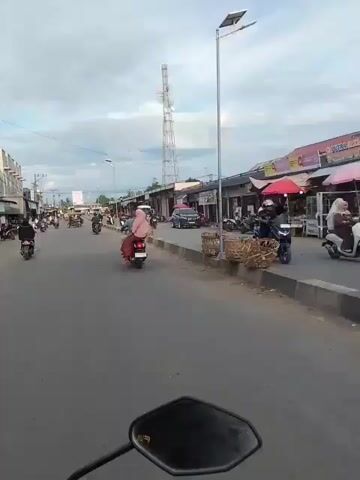 A first-person perspective video shows a street scene. A motorcycle is moving down a paved road. On either side of the road are buildings that appear to be shops or stalls, some with awnings. Several people are on motorcycles, either driving or as passengers. A tall cell tower is visible in the distance. The sky is cloudy.