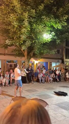 A male street performer plays a saxophone for an audience seated on benches. The performer wears a white t-shirt and blue shorts and stands in a paved plaza under a large tree. The audience is comprised of men, women, and children of various ages. Behind the audience are shops and buildings. The scene is lit by streetlights and the setting sun, creating a warm, evening atmosphere.