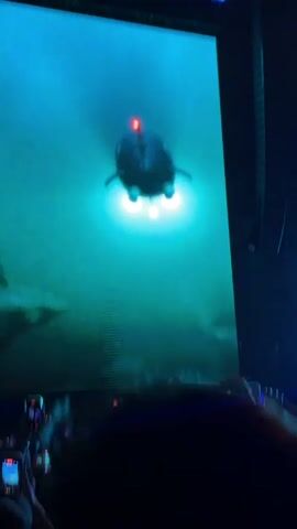 A concert stage with a large screen displaying a blurry image of what appears to be a helicopter against a green and blue background. Lights on the stage are blue and white, and there is smoke or haze. In the foreground, the silhouettes of concertgoers are visible, many holding up their phones to record. The text "PARK" is visible on the screen to the right of the helicopter image.