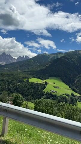 This video captures a scenic view of a mountainous landscape in St. Peter - San Pietro, Italy. The foreground features a guardrail, suggesting the perspective is from a vehicle or a roadside viewpoint. Beyond the guardrail, lush green grass dotted with white wildflowers grows on a hillside.

The midground is dominated by dense, dark green pine forests covering the slopes of rolling mountains. Patches of bright green meadows are visible amidst the trees, with small clusters of houses nestled within them, indicating a rural settlement. The houses appear to have red-tiled roofs, typical of the region.

In the background, majestic, jagged mountain peaks rise against a bright blue sky. The mountains have a rocky, dramatic appearance. The sky is a vibrant blue with scattered, fluffy white clouds. The lighting suggests it is daytime, likely midday or early afternoon, given the brightness and the shadows cast.

The overall atmosphere is serene and picturesque, showcasing the natural beauty of the Italian Alps. There are no people or specific objects of interest beyond the natural scenery and the man-made guardrail. The video appears to be a static shot, offering a still panorama of the breathtaking landscape.