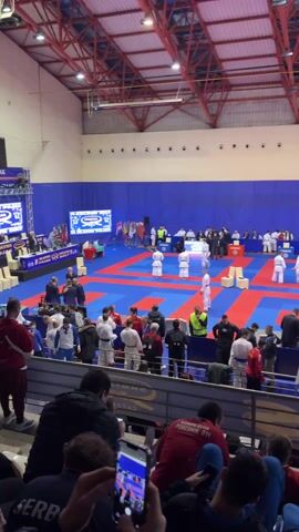 The video shows a martial arts tournament taking place indoors. Multiple red and blue mats are arranged side-by-side, each holding two competitors dressed in white uniforms. Referees and judges stand around the mats, observing the matches. A crowd of spectators fills the bleachers, some holding up phones to record the event. Banners and flags representing different countries are visible, and the building has a high ceiling with a red metal framework.