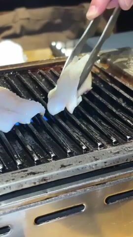 This is a close-up shot of food being grilled on a metal grate. There are two pieces of white, raw food on the grate, and a third piece is being held by silver tongs and lowered onto the grate. Blue flames are visible beneath the grate, indicating that the grill is on. The background is dark and out of focus. The focus is on the food and the grilling process.