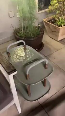 The video shows an outdoor patio scene at night. It is raining, and a two-tiered gray serving cart has collected a significant amount of water on both levels. Potted plants are visible in the background. The camera briefly pans upward to show the rest of the patio, then focuses on a person with long, wavy brown hair, wearing a gray t-shirt, who is looking directly at the camera with a slightly quizzical expression. The person appears to be standing on the patio, possibly observing the rain.