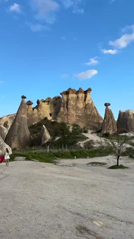 This video captures a scenic tourist experience in Çavuşin Köyü, Cappadocia, Turkey. The foreground features a dirt ground and road where several people are gathered. A red vintage convertible car is parked, and people are seen posing with it and a white horse ridden by an individual. In the background and midground, distinctive "fairy chimney" rock formations, characteristic of Cappadocia, rise dramatically against a clear blue sky with scattered white clouds. Some sparse green vegetation, including small trees and grass, is visible among the rock formations and on the ground. People are casually walking and standing, appearing to be tourists enjoying the unique landscape. The red car's license plate "50 AB 269" is visible. The scene depicts a sunny, pleasant day.