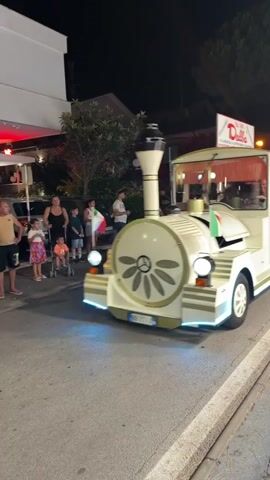 A white, train-like vehicle is driving down a street at night. The vehicle has a vintage design with gold trim and decorative elements. It has an open passenger area with people sitting inside, some of whom are children. An Italian flag is attached to the front of the vehicle. There are businesses on the side of the street, one of which has a red sign that says "Dolciaria Dalba" and "dal 1930". Another sign behind it says "La Capricciosa". People are standing on the sidewalk watching the vehicle. The street is paved and there are lights illuminating the scene. The video captures the vehicle from a side angle as it moves forward.