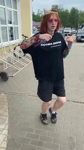 This is a video clip of a young person with ginger hair, wearing sunglasses, a black t-shirt with white text, black shorts, a baseball cap, and black sneakers. The t-shirt has the Russian text "Хорошие девочки (Good girls) Не попадают в историю (Don't make history)". The person has colorful tattoos on their arms and is holding a purple object, possibly a chocolate bar, in their right hand. They are dancing or posing in a lively manner, with their body bent and one leg extended. The setting appears to be an outdoor paved area, possibly a sidewalk or plaza, with a building to the left and a row of parked cars in the background. A bicycle is also visible on a bike rack. The sky is overcast, suggesting a cloudy day. The person's expression and posture convey a sense of confidence and playfulness. The location context provided is Bezhetsk, Russia.