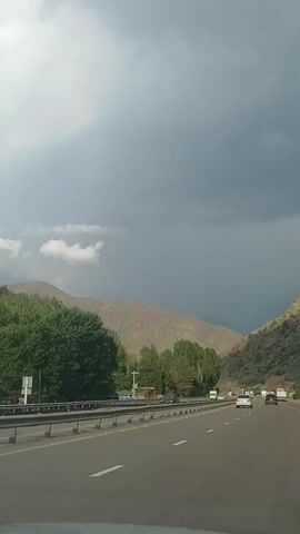 A vertical, first-person perspective video captures a highway scene with a dramatic sky. The highway appears to be a multi-lane road with a metal guardrail separating it from the left shoulder. Cars and trucks are visible on the road, traveling in the same direction as the camera. Lush green trees line the left side of the highway, and beyond them, mountains rise under a turbulent, overcast sky. The clouds are dark and heavy, suggesting impending rain or a storm. The overall environment appears to be a natural landscape adjacent to a developed transportation route.