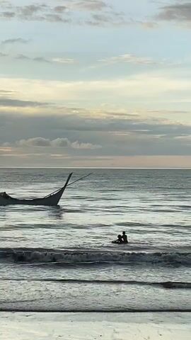 The video shows a serene beach scene at dusk. In the foreground, gentle waves lap against the shore. Further out in the ocean, a small, traditional boat, possibly a fishing vessel, is partially submerged, listing to one side with its mast tilted. In the middle ground, two individuals are in the water, one sitting and the other appearing to be swimming or wading. They are positioned between the shore and the partially submerged boat, indicating they are enjoying the water or perhaps interacting with the boat. The sky above is filled with soft, muted clouds, tinged with the warm hues of a setting sun, creating a peaceful and tranquil atmosphere.