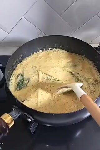 A close-up, top-down shot shows a black, non-stick frying pan filled with a bubbling, creamy yellow liquid and several large green leaves. The pan is situated on a black stovetop. A person's hand, holding a spatula with a white silicone head and a light-colored wooden handle, is actively stirring the contents of the pan. The spatula is angled, and its head is submerged in the liquid, gently moving it around. The background features a white tiled wall with a subtle horizontal grout pattern. The overall impression is that of food being cooked, likely a curry or sauce, given the creamy texture and leafy ingredients.