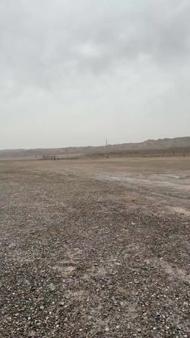 The video shows a barren, gravel-covered landscape under a very overcast sky. Low, dry hills are visible in the distance. Power lines stretch across the scene. The wind seems to be strong, indicated by the sound. The camera pans to show several vehicles parked in an area with a low, rectangular concrete structure, possibly an entry point to an underground area. No people are visible.