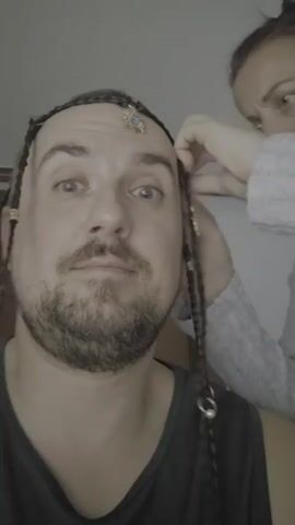 A close-up, shaky, first-person perspective video shows a man with graying hair braided into several thin braids adorned with gold beads and rings. A small, ornate gold and blue jewel is attached to his forehead. A woman is visible in the background, to the right, with her hands near the man's head, seemingly adjusting his hair. The man makes various facial expressions, including surprise, a questioning look, and a pouting grimace. The environment appears to be indoors, with a neutral-colored wall in the background.