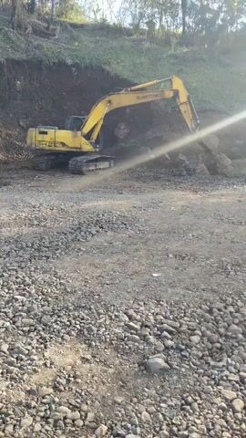 A yellow excavator is positioned on a gravel surface, its boom extended and bucket digging into a pile of rocks and dirt. The excavator appears to be on a construction site or excavation area, with a steep, vegetated embankment to its left. Sunlight is casting a glare across the scene.