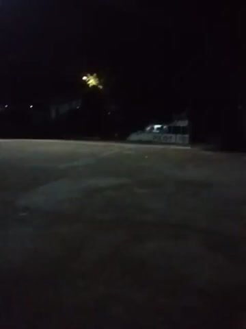 The video shows a dark scene at night. A building with the word "PILOT" on it is visible in the foreground, towards the left side. In the background, across what appears to be a body of water, there's a structure with lights reflecting on the water. The lights suggest the presence of a bridge or a building illuminated at night. The right side of the view is primarily dark, possibly a wall or ground surface.