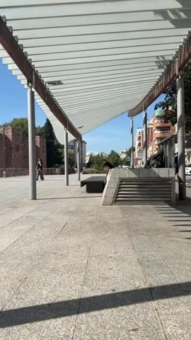 A young man with short brown hair, wearing a black t-shirt with a circular graphic on the back and loose brown cargo pants, is skateboarding under a modern, white-slatted overhang supported by grey cylindrical poles. He approaches a dark-colored bench positioned at an angle. As he reaches the bench, he ollies over it. He lands on the other side of the bench with his skateboard. He then turns and walks away from the bench, facing the camera. The environment is an urban plaza with a patterned stone ground and a blue sky with scattered clouds. To the left, there is a brick building and some trees. To the right, there are modern buildings and streetlights. Another person is visible in the background, walking further down the plaza.