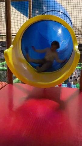 A young boy with blond hair slides down a yellow and blue enclosed slide in an indoor play area. He is wearing a light-colored sweatshirt and gray sweatpants with socks. He lands on a red padded surface at the bottom of the slide, falls over, and then smiles at the camera. The play area is enclosed with netting and has a green and black floor.
