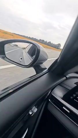 The video shows a view from inside a car traveling on a road. The camera is looking out the side of the car, capturing the passing scenery and the side mirror. The scenery consists of a field of golden crops, a grey road with white lines, and a cloudy sky. The car appears to be in motion, as indicated by the blurred road and the movement of objects in the distance. There are no people visible in the video. The perspective is unusual, with the car and the road appearing tilted or even upside down at times, suggesting a possible creative filming technique or an issue with camera orientation.
